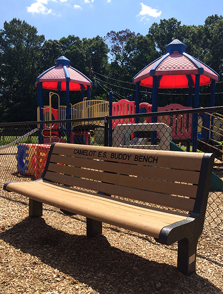 Color photograph of the Buddy Bench. Playground equipment is visible behind the bench. 