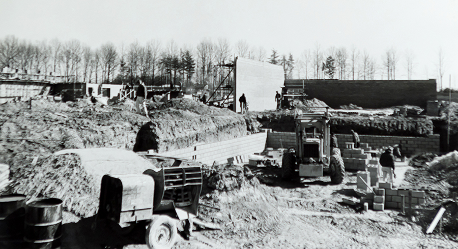 Black and white photograph of Camelot Elementary School taken in 1968 during construction of the building. Construction vehicles and supplies can be seen in the foreground. The cinderblock walls are starting to be erected. In the distance, tall trees without any leaves can be seen. Construction workers can be seen in different places around the site, working on the masonry. 