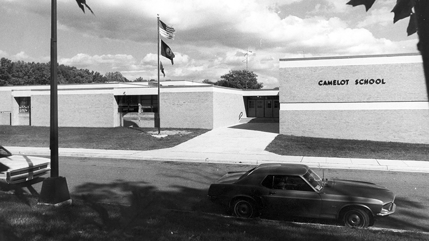Black and white photograph of the front exterior of Camelot Elementary School taken in the 1970s. The main entrance doors and flagpole are visible. Two cars are parked in front of the school. There are no bushes or shrubs in front of the building, so this picture may date from the earliest years after Camelot opened.