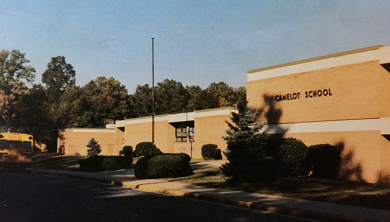 Color photograph of the front exterior of Camelot Elementary School from our 1981 to 1982 yearbook. Several large shrubs and a pair of evergreen trees have been planted in front of the school. A school bus can be seen on the far left, parked in front of the building. 