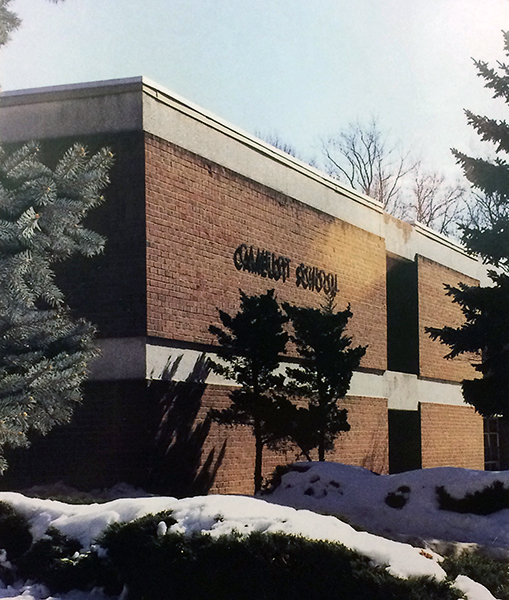 Color photograph of the front exterior of Camelot Elementary School from our 1999 to 2000 yearbook. Two large evergreen trees can be seen on the left and right side of the picture. In the center, the shrubs in front of the school are covered with snow. The sun is shining on the side of the building, casting long shadows. 
