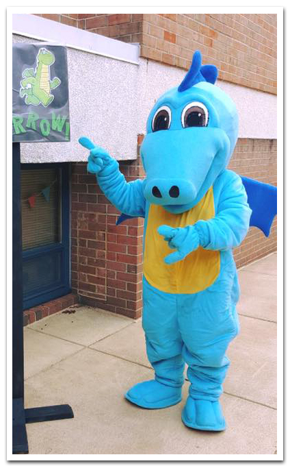 Photograph of an unknown person wearing the Flames mascot costume. The dragon costume has light blue fabric on the arms, legs, and head, dark blue fabric on the wings, and gold fabric on the belly. The mascot is standing on the sidewalk in front of our school pointing to an illustration of Flames on a sign. 