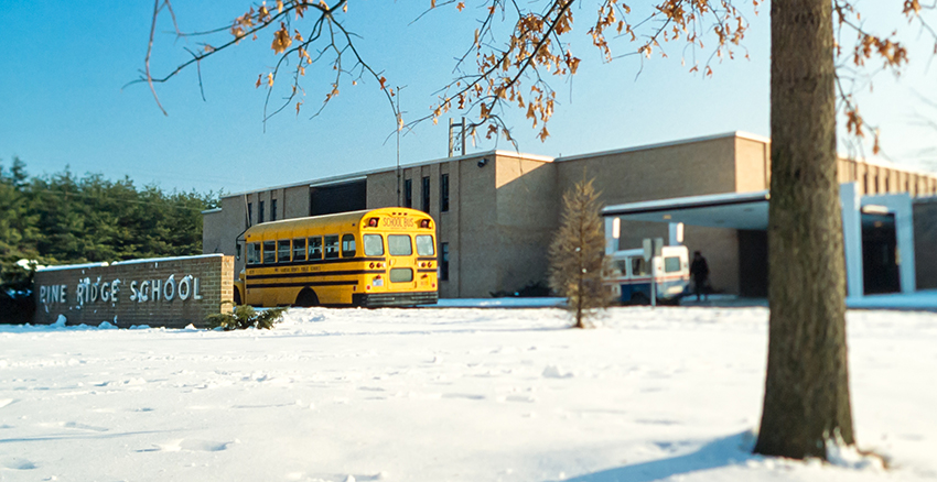 Color photograph of the front exterior of Pine Ridge Elementary School taken around 1980. A school bus and U.S. postal truck are parked in front of the building. There is snow on the ground and the trees in the foreground have lost most of their leaves. 